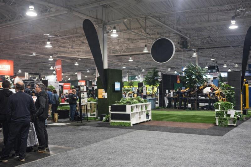 Indoor trade show exhibition featuring synthetic turf display booth with landscaping setup and visitors exploring products